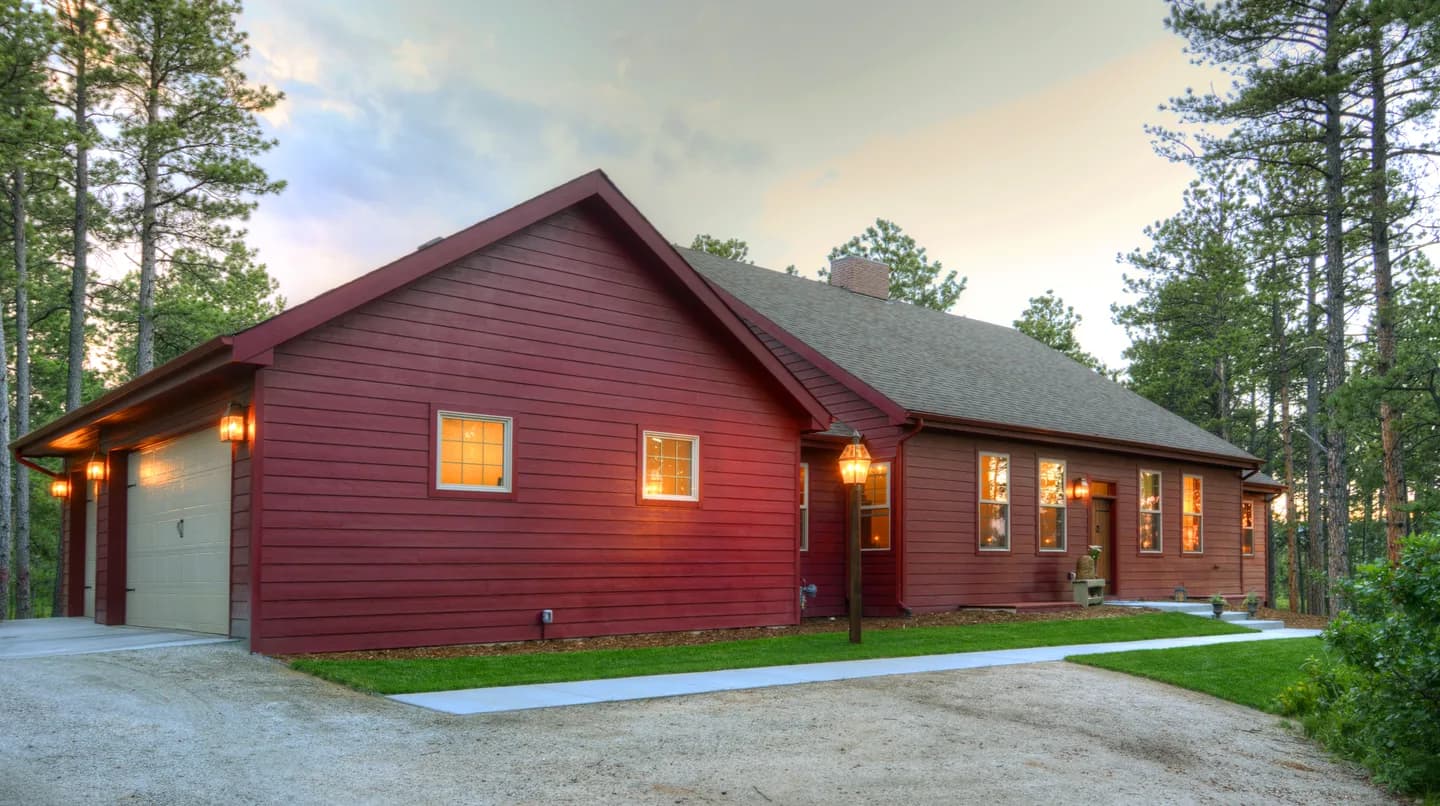 Barn-red custom ranch home at dusk with attached garage, warm exterior lighting and pine forest surroundings