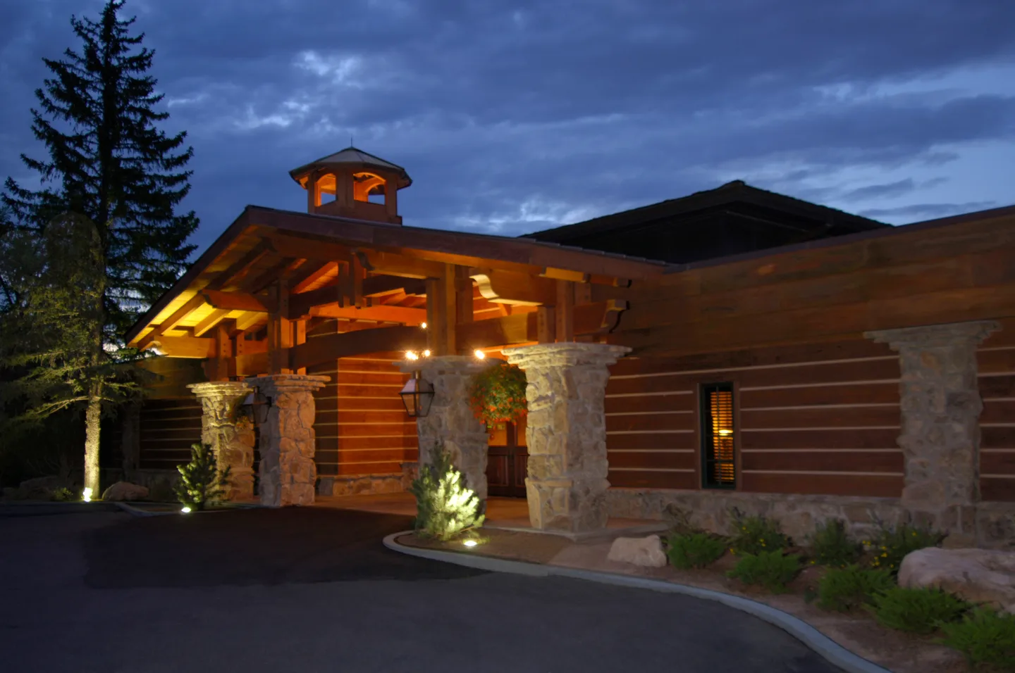 Remodeled lodge entry at dusk with timber porte-cochere, cupola, stone columns and uplit landscaping