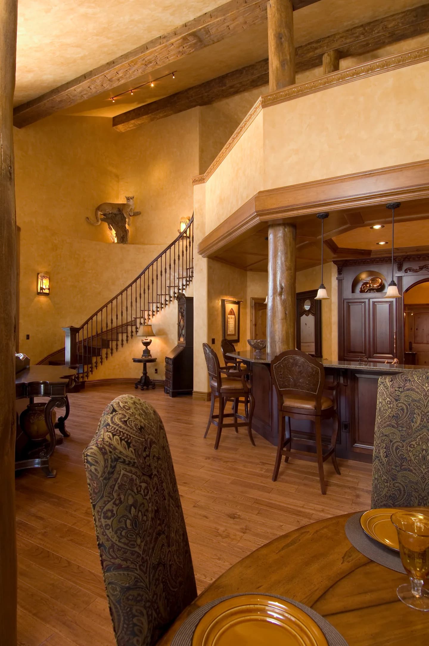 Custom wet bar with leather barstools beside a curved iron-railed staircase in a two-story timbered space