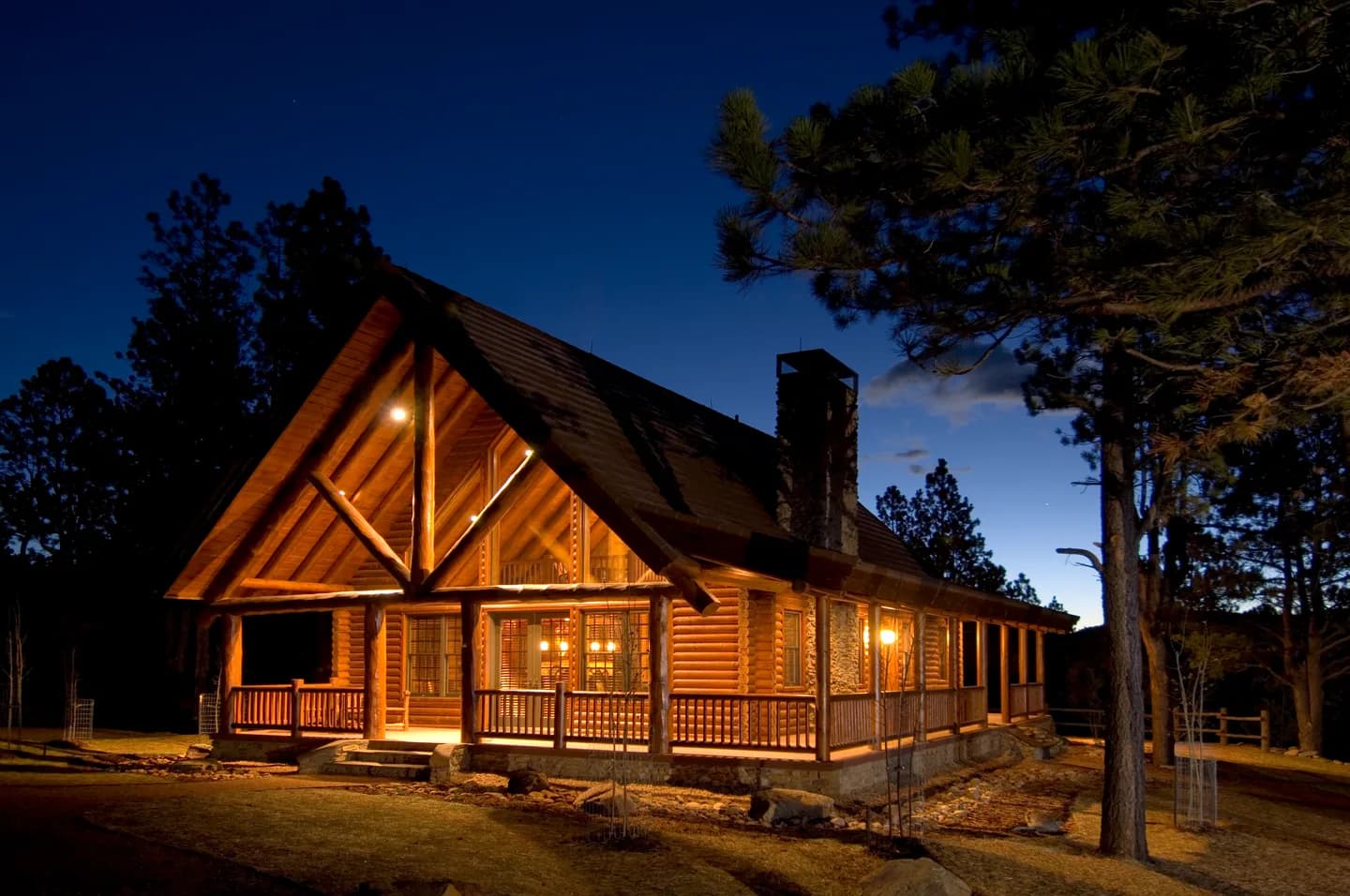 Log home glowing at twilight with wraparound porch, steep gable and stone chimney among ponderosa pines
