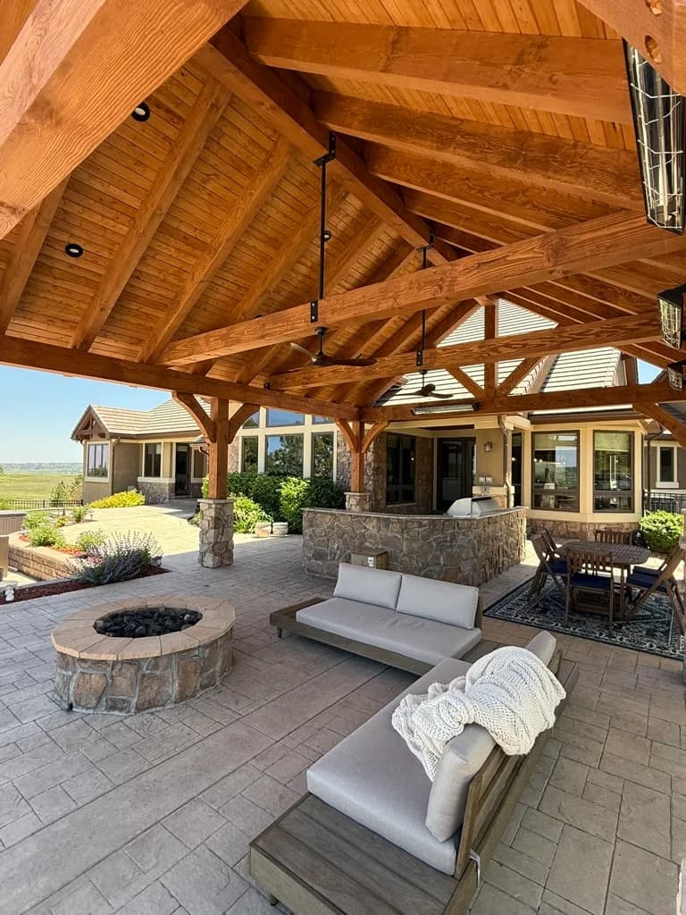 Timber pavilion with exposed trusses sheltering a patio with stone fire pit and outdoor sectional seating