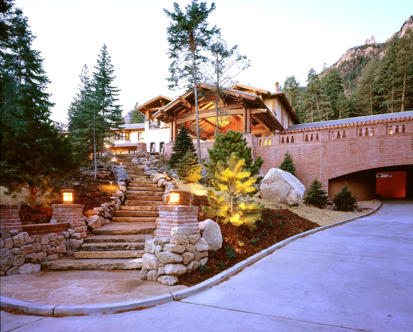 Estate entry at dusk with granite stair, lit brick piers, timber great room gable and arched garage tunnel