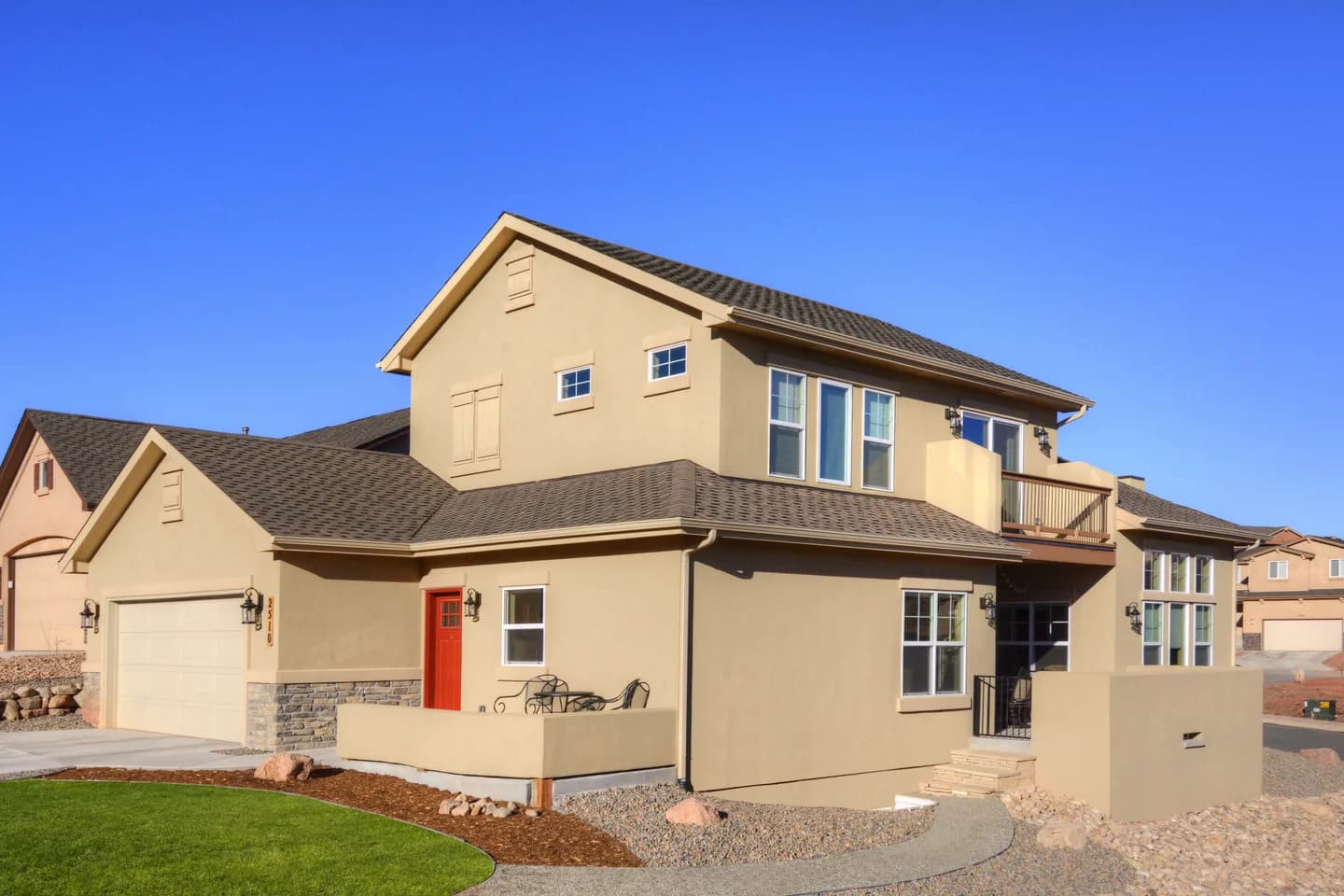 Two-story stucco custom home with covered balcony, red front door and attached garage under blue sky.